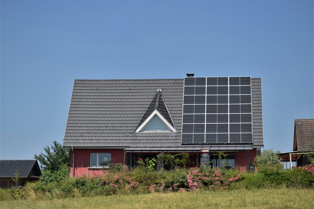 A red-roofed house with solar panels on the roof sits in a green, grassy field with bushes and trees around it. The sky above is blue with some clouds.