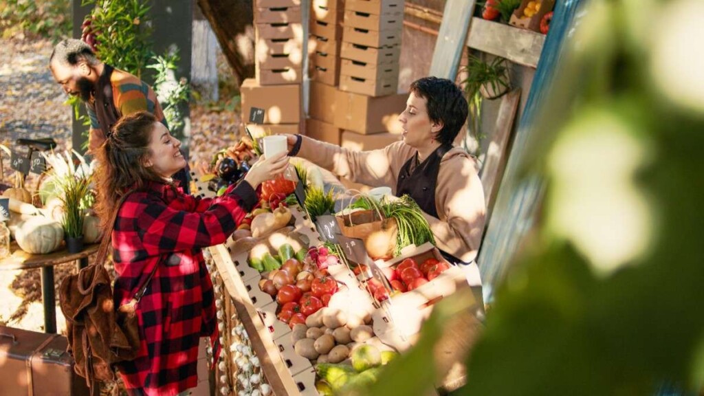 A friendly vendor at a farmers' market stall hands a cup to a customer surrounded by fresh organic produce.