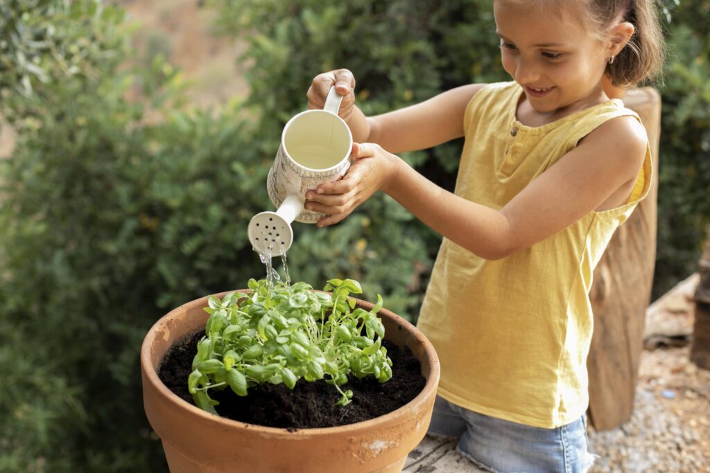 A smiling young girl in a yellow tank top uses a small white watering can to water a freshly potted green plant in a terracotta pot outdoors.