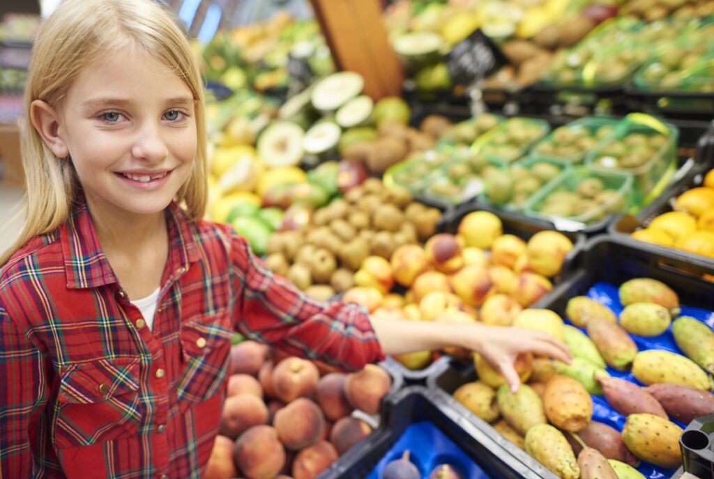 A smiling young girl with blonde hair and a red plaid shirt looks at the camera while reaching for prickly pear fruits in a bin at a brightly lit grocery store produce section.