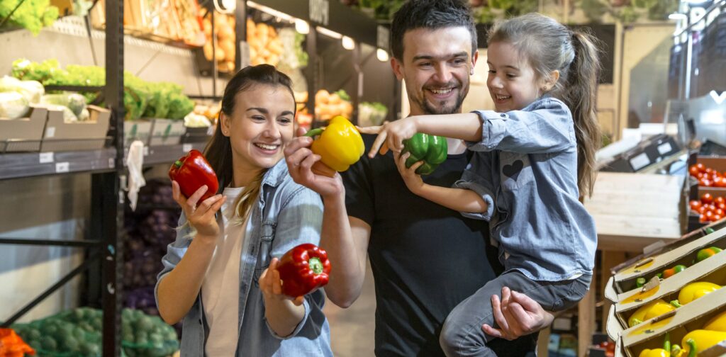 h A smiling family of three—a mother, father, and young daughter—happily chooses colorful bell peppers together in the produce aisle of a supermarket.
