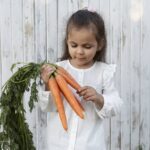 A young girl with dark hair in a white shirt looks down at a bunch of bright orange carrots with green tops that she is holding in her hands, against a white wooden fence background.